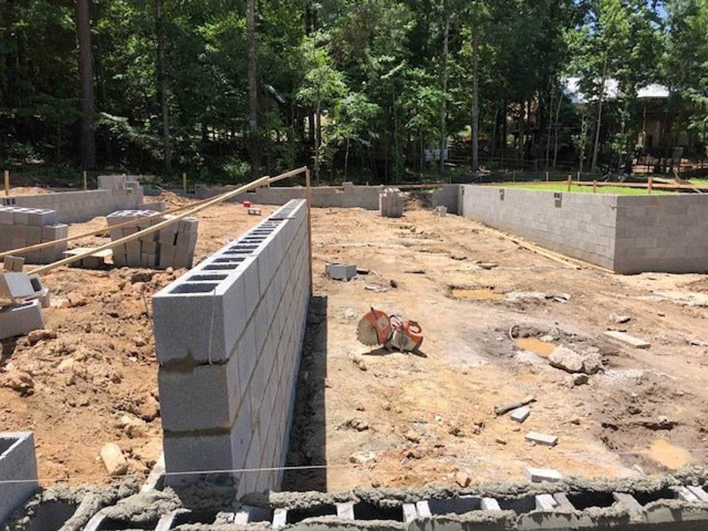 Brick wall under construction beside wooden fence, surrounded by soil and rocks, with trees in background.