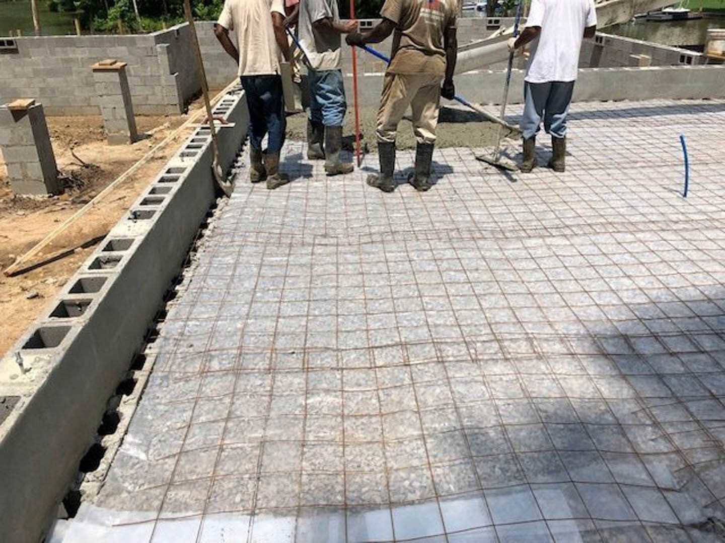 Men standing on unfinished concrete floor outside brick home, one holding a shovel, another with a cane, surrounded by trees and sidewalk, wearing casual clothing and jeans