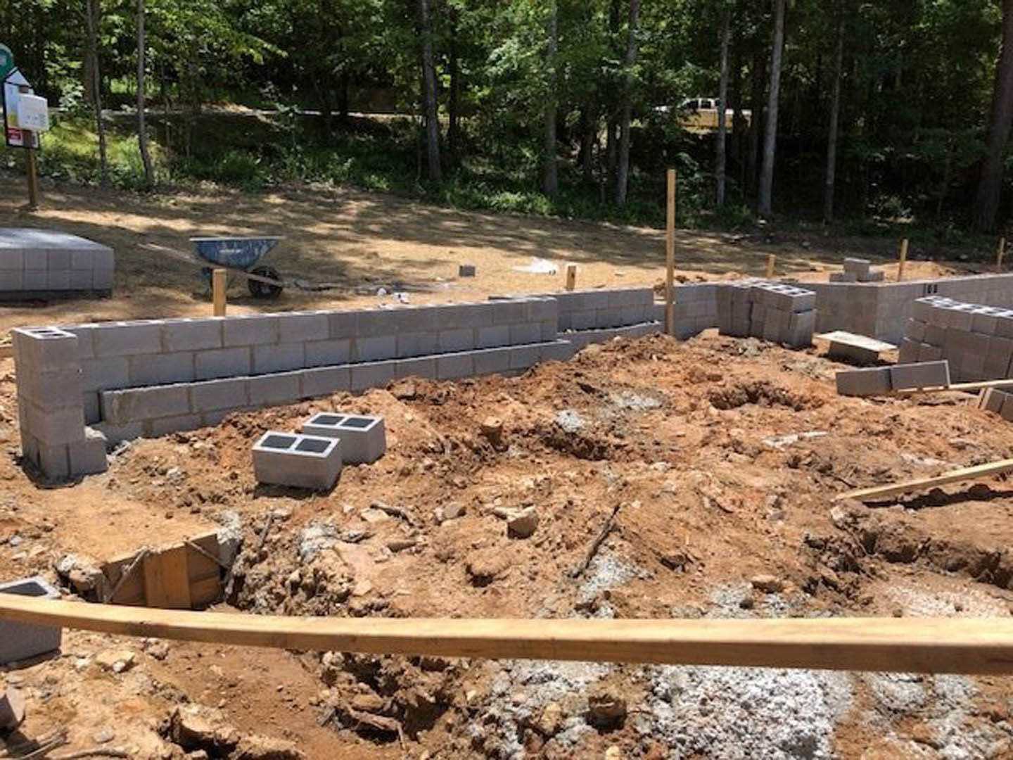 Bricks stacked on bare soil beside a vertical wooden pole at a residential construction site, surrounded by scattered rocks and trees in the background