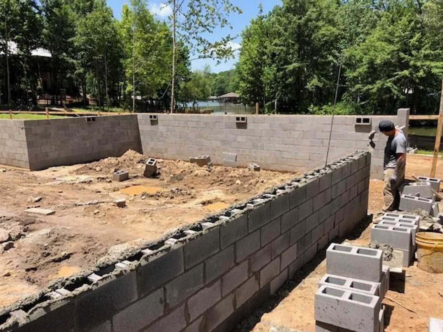 Man standing in front of partially built brick wall and foundation at residential construction site, trees and lake in background