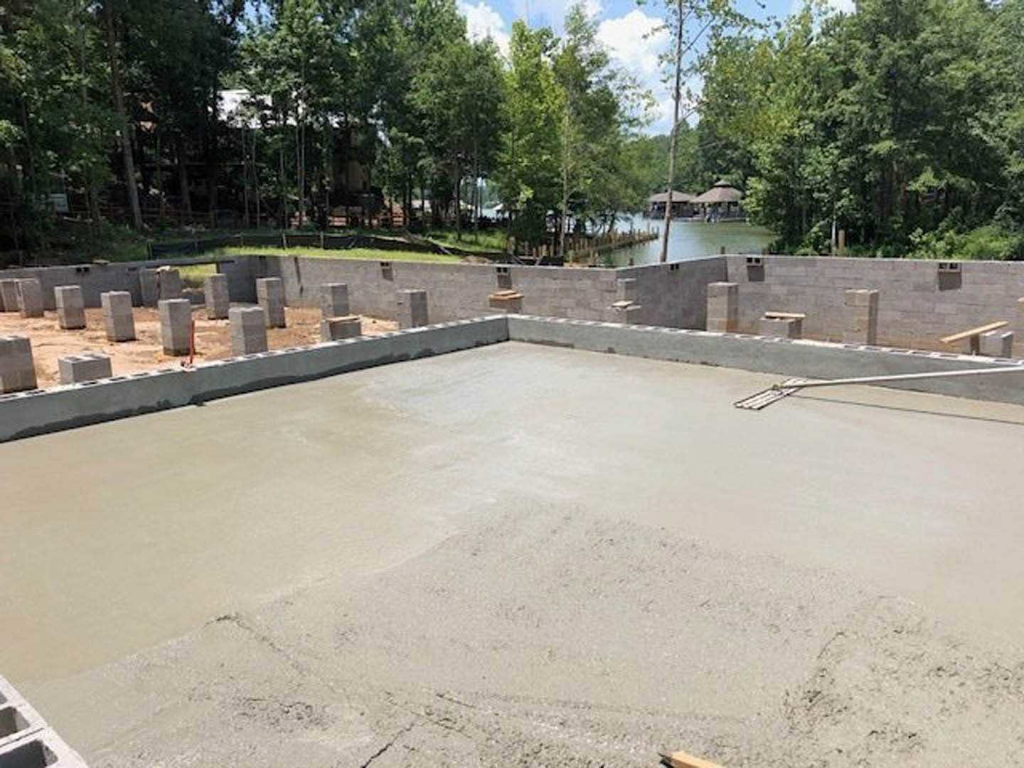 Freshly poured concrete foundation slab with wooden formwork, surrounded by dirt and construction materials, trees and a lake visible in the background.