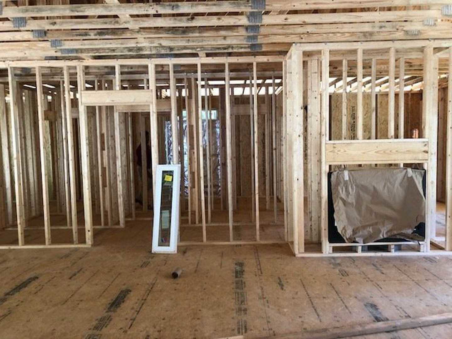 Exposed wooden framing and beams inside a house under construction, unfinished wood floor with a roll of cardboard, white insulation sheet draped over a section, pallet and