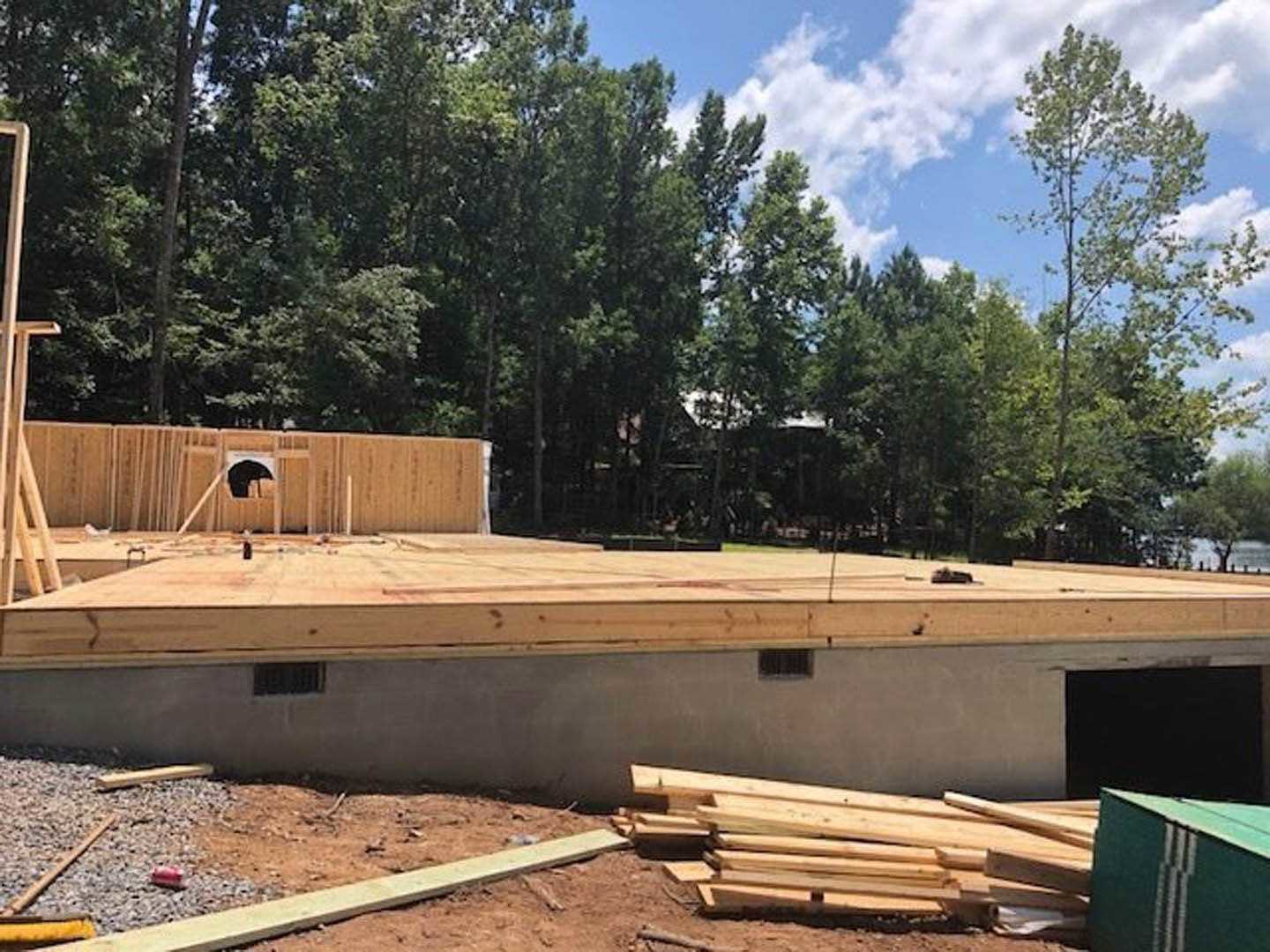 Framed wooden structure under construction, stacks of lumber on bare ground, surrounding trees and open sky in background