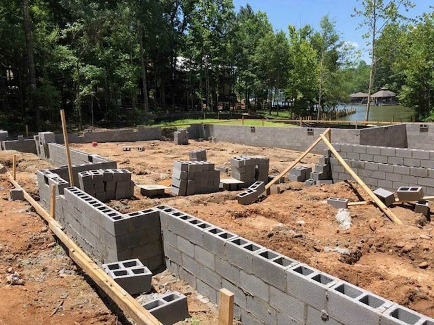 Concrete foundation blocks set on soil at a residential construction site, surrounded by trees under a partly cloudy blue sky