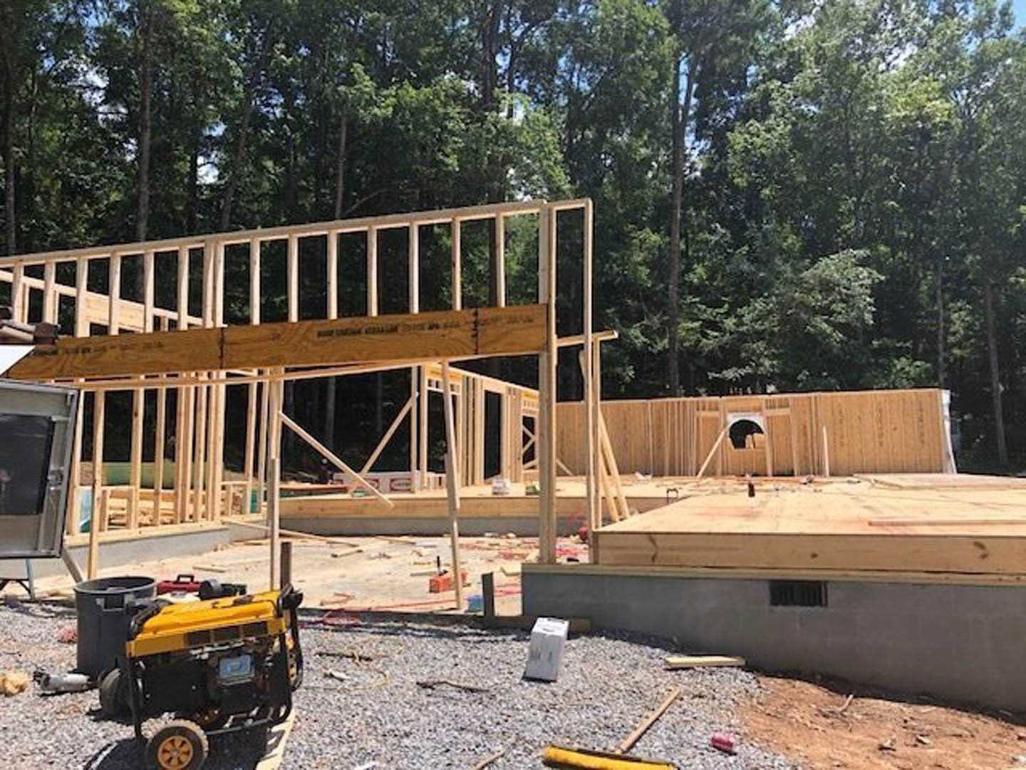 Wooden framing for a custom home under construction, white concrete block on ground, yellow generator on gravel driveway, trees in background