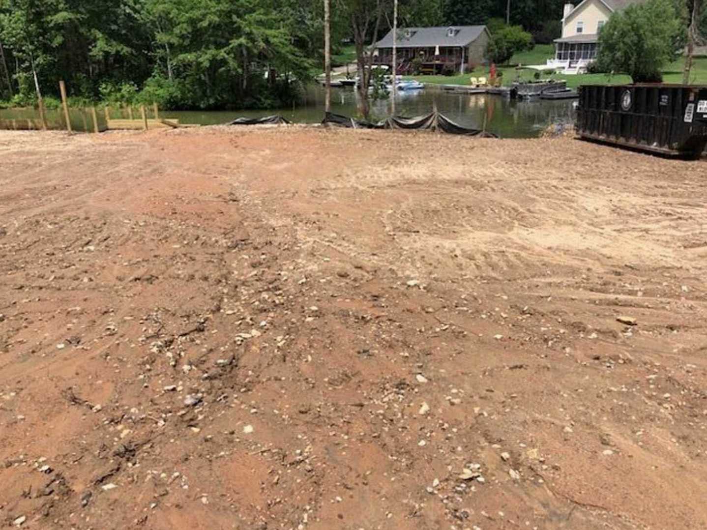 Brown dirt lot beside a small pond, bordered by trees and several houses with grey roofs in the background