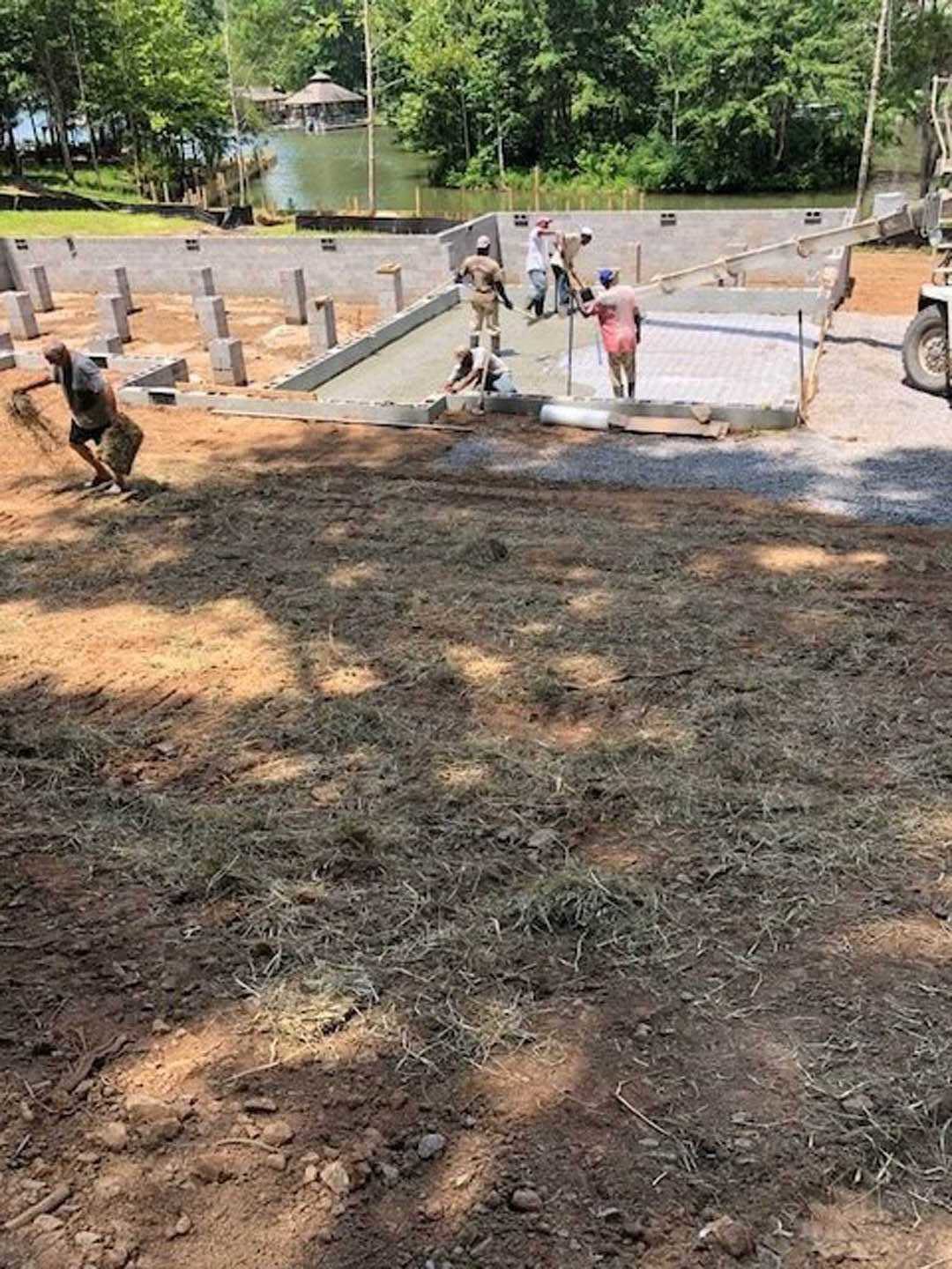 Men assembling framing and working with construction materials on a dirt lot surrounded by grass, trees, and playground equipment