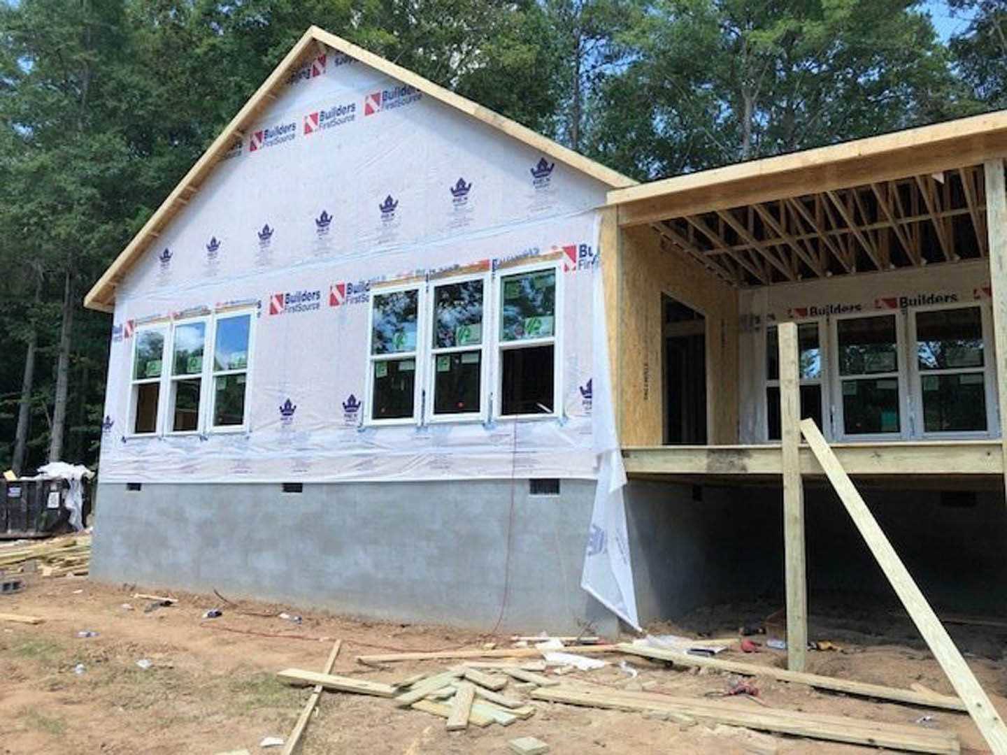 Wood-framed house under construction with exposed lumber, partially built exterior walls, scattered wood piles, and workers on site