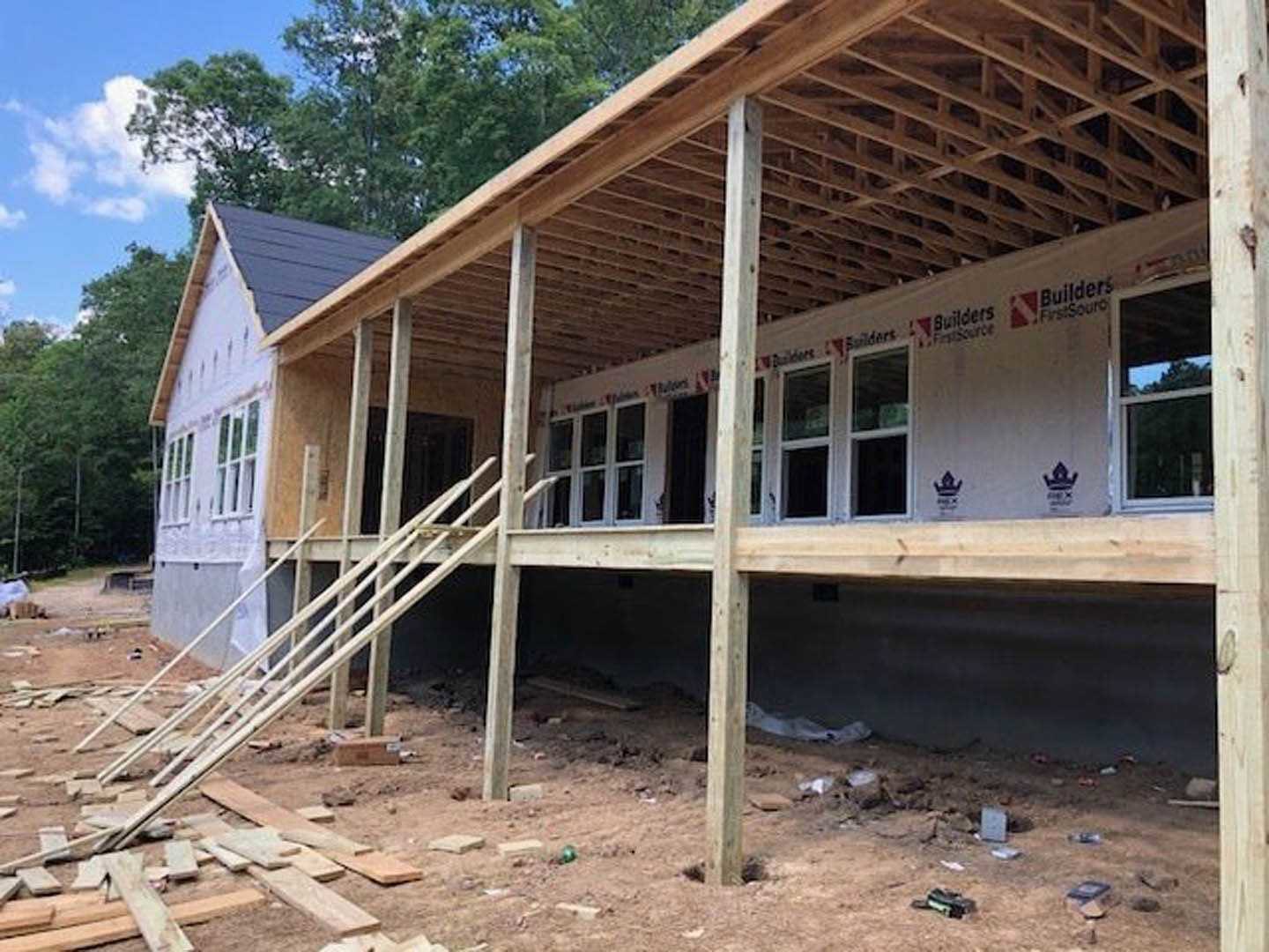Framed house under construction with exposed lumber, wooden ladder leaning against unfinished wall, white-framed window overlooking trees, logo visible on white surface