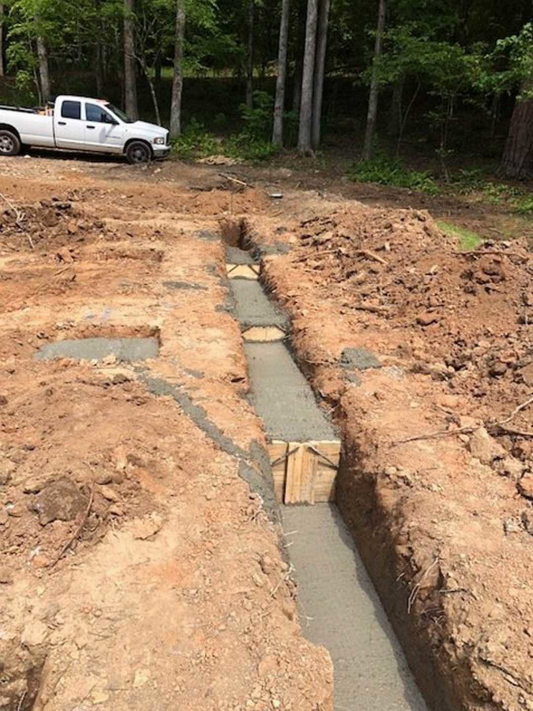 Dirt trench with exposed concrete and wood, white truck parked behind, surrounded by trees and forest vegetation