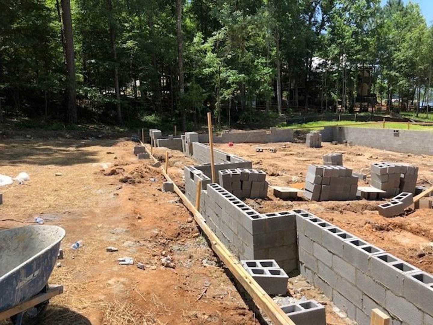 Concrete foundation wall and stacked bricks on a residential construction site, with exposed soil and scattered building materials, surrounded by trees.