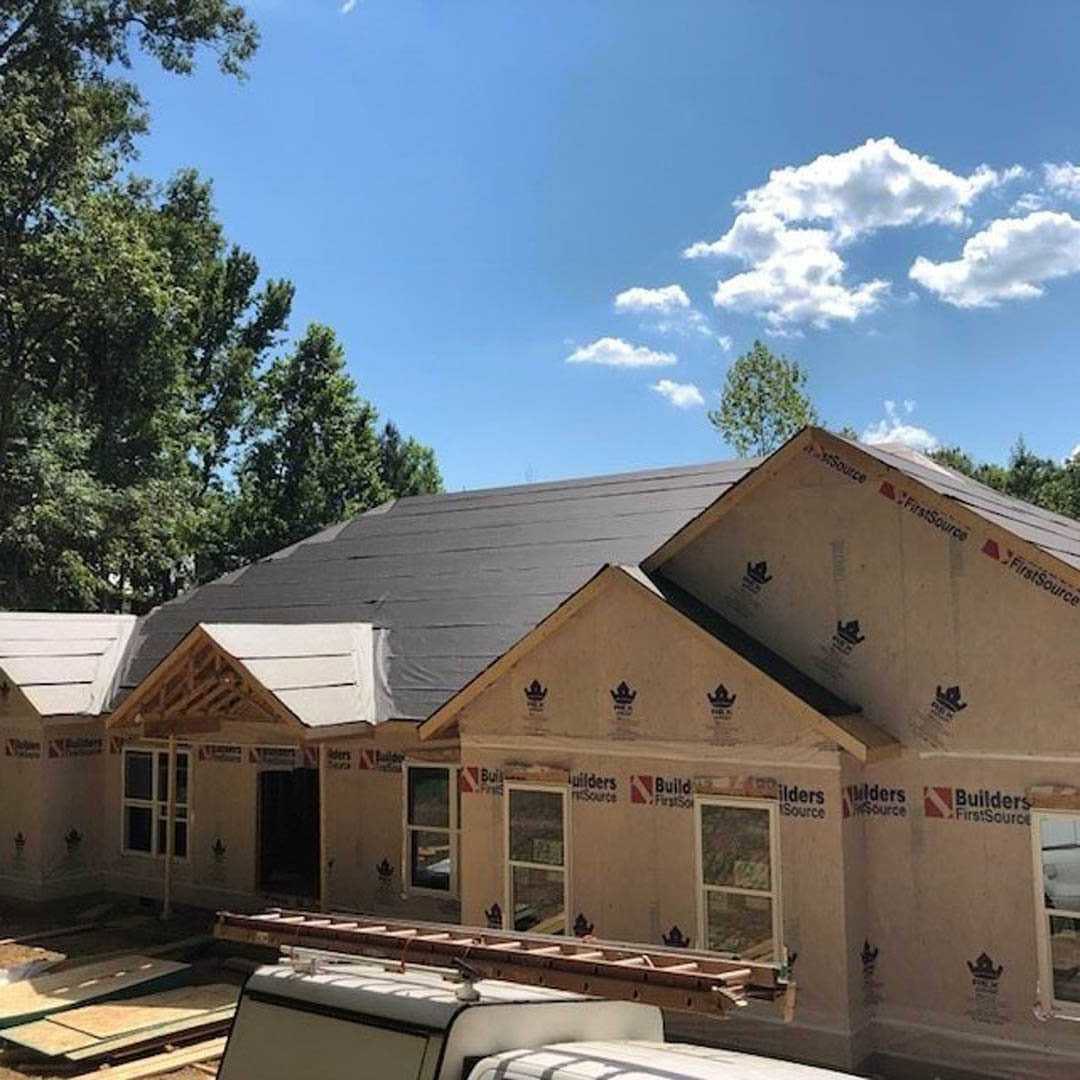Framed house under construction with exposed plywood, parked pickup truck in driveway, mature trees surrounding site, cloudy sky overhead