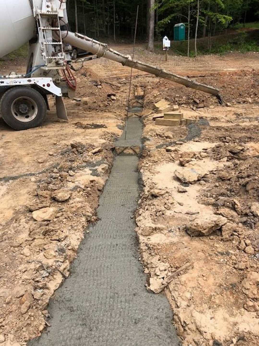 Concrete mixer pouring fresh concrete into a dirt trench, green container with white top nearby, tire and wheel of vehicle visible, machine with long pipe, concrete drainage ditch