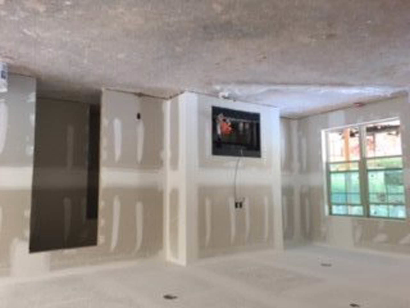 Living room with mounted flat-screen TV on a plaster wall, large window with natural light, neutral carpet flooring, and white ceiling.