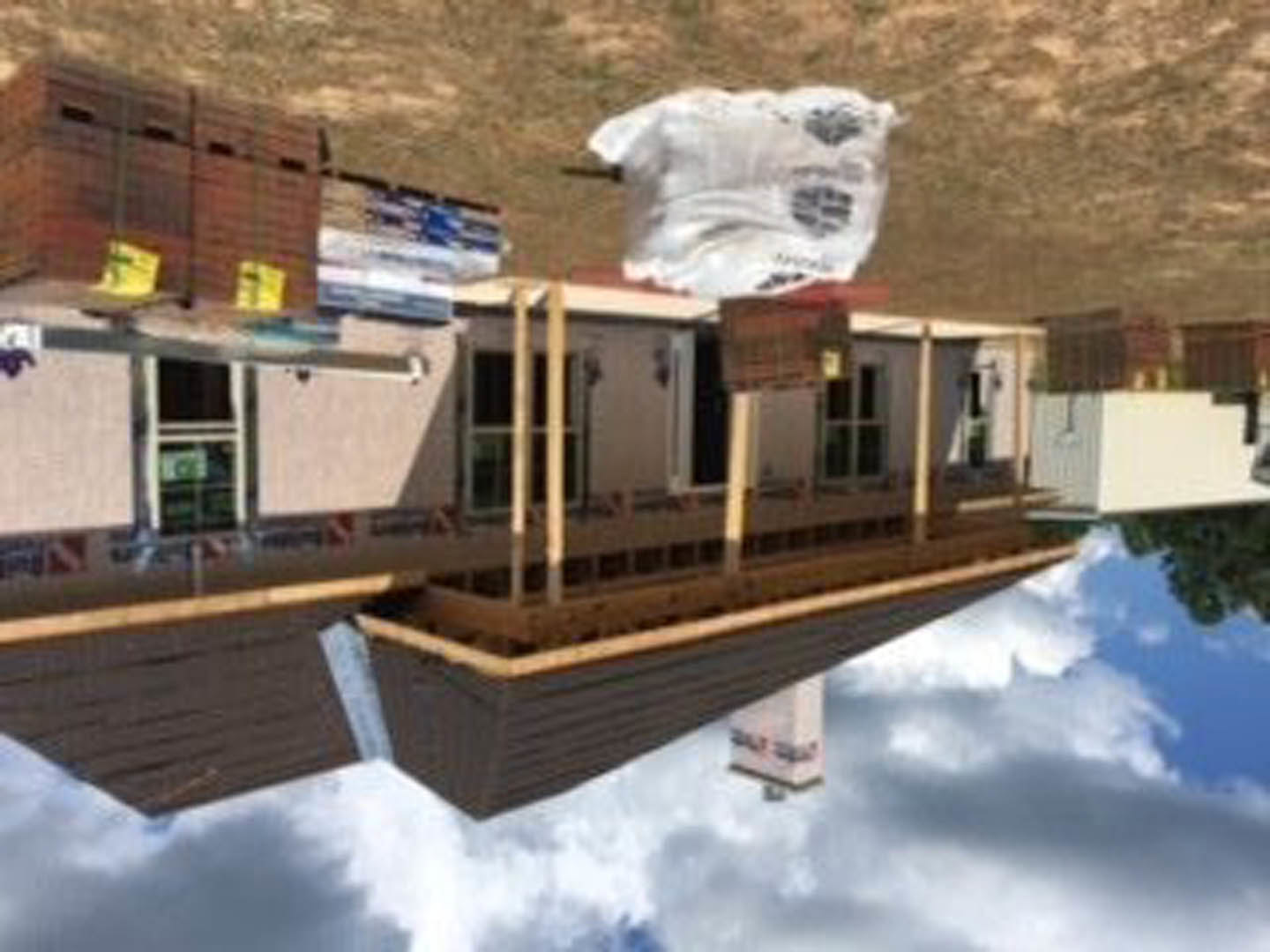 Framed house under construction with exposed wood beams, white bag of materials on dirt ground, cloudy sky overhead