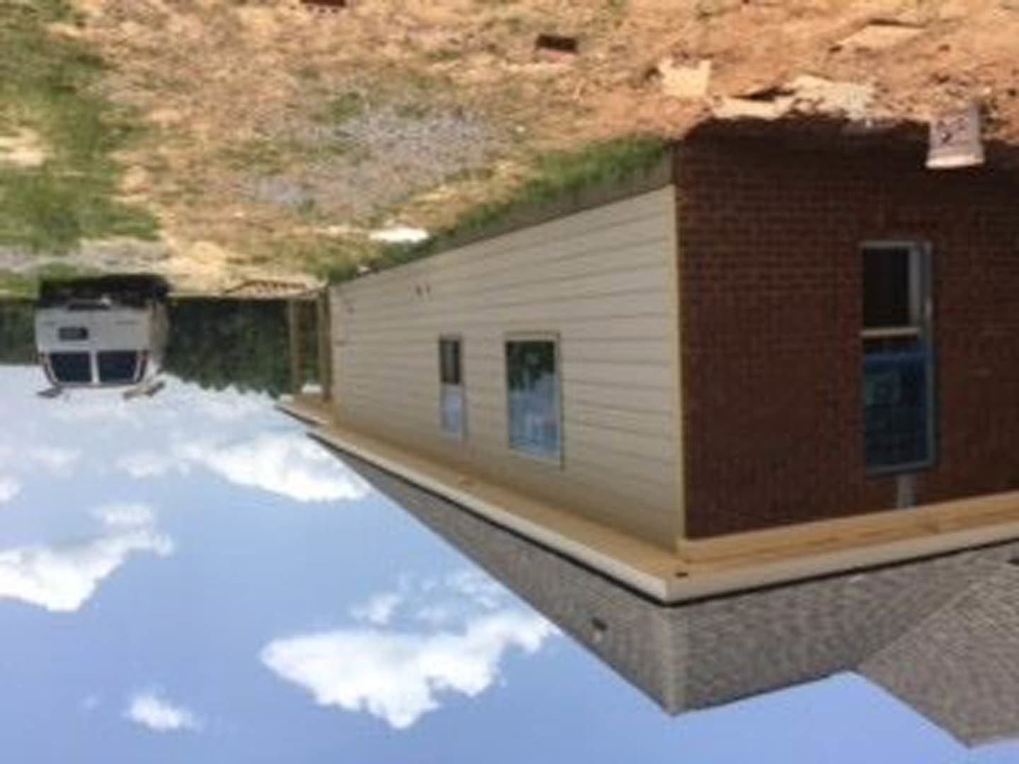 Modern home exterior with large windows reflecting blue sky and clouds, flat roof with green grass, landscaped ground, and contemporary architectural lines