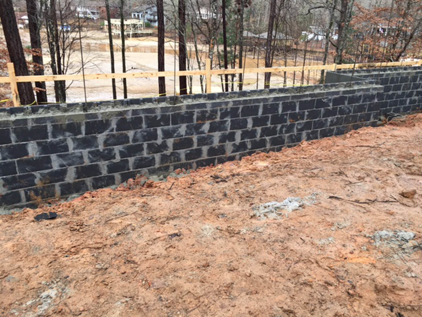 Red brick wall with dirt at the base, wooden fence and trees in the background
