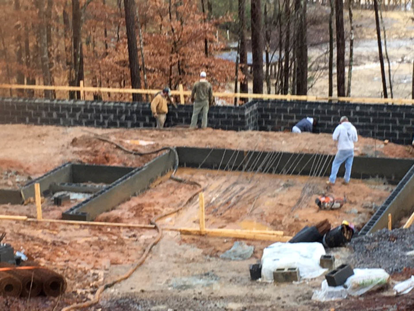 Workers assembling foundation on construction site, surrounded by dirt, trees, and building materials