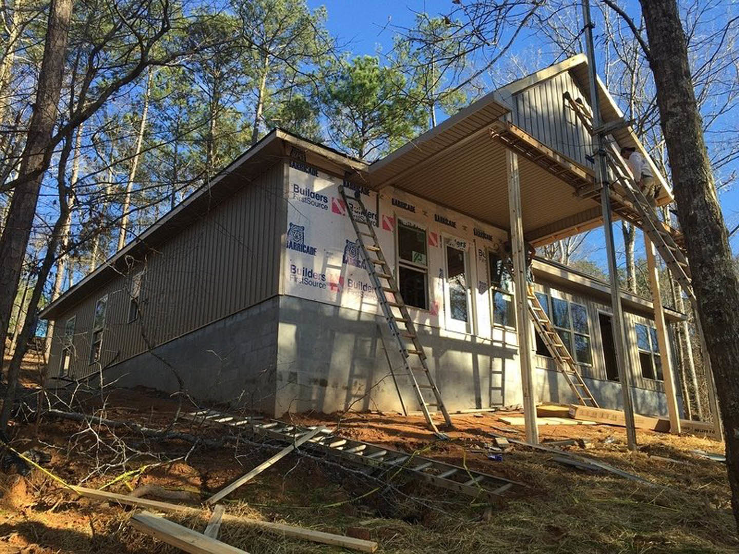 Two-story house under construction with exposed framing, ladders propped against exterior walls, large windows, and mature trees surrounding the site