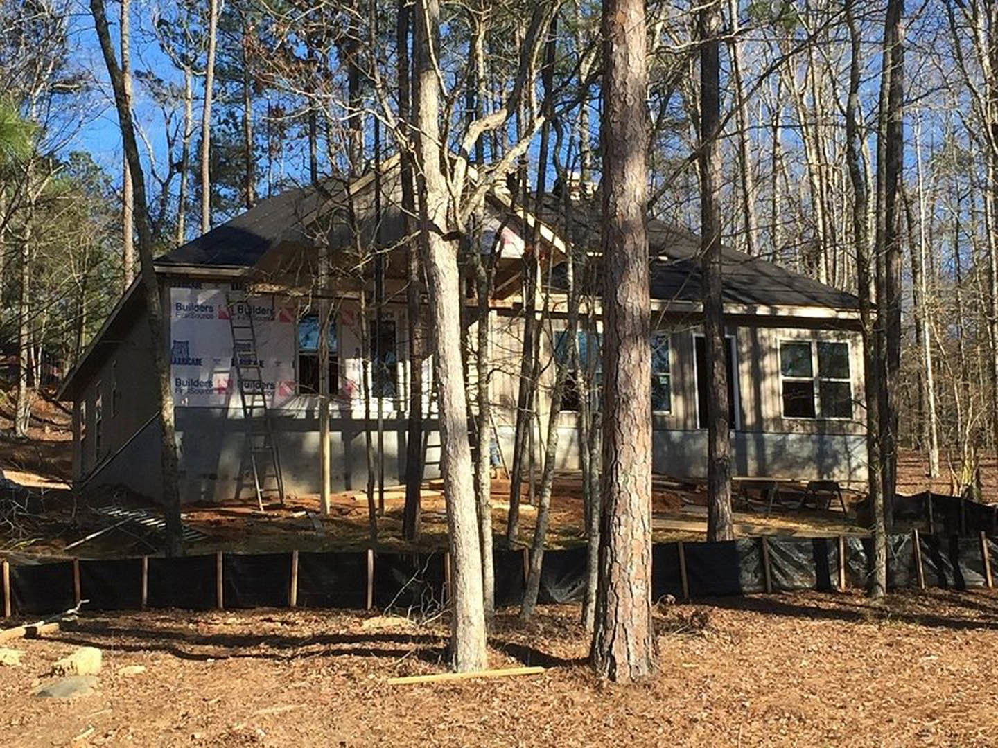 Wood-framed house under construction surrounded by tall trees, white window frames visible, unfinished exterior walls, forested background