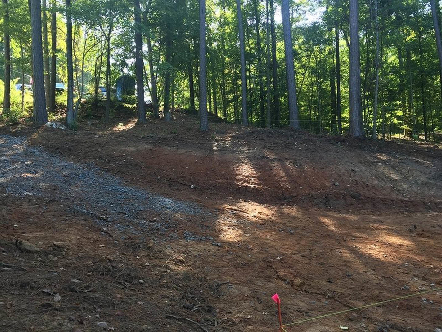 Dirt hill bordered by a grove of trees, rocky trail in foreground, red flag on pole, rope and blue object partially visible
