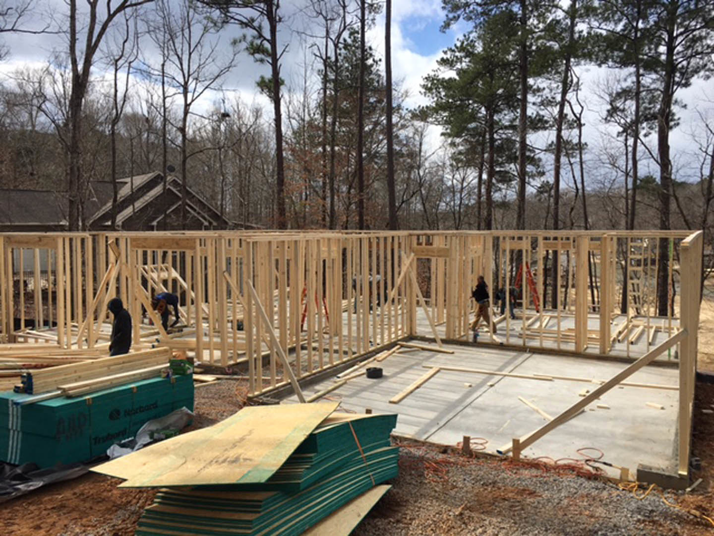 Framed wooden house under construction with workers, stacks of plywood, blue toolbox, and surrounding trees