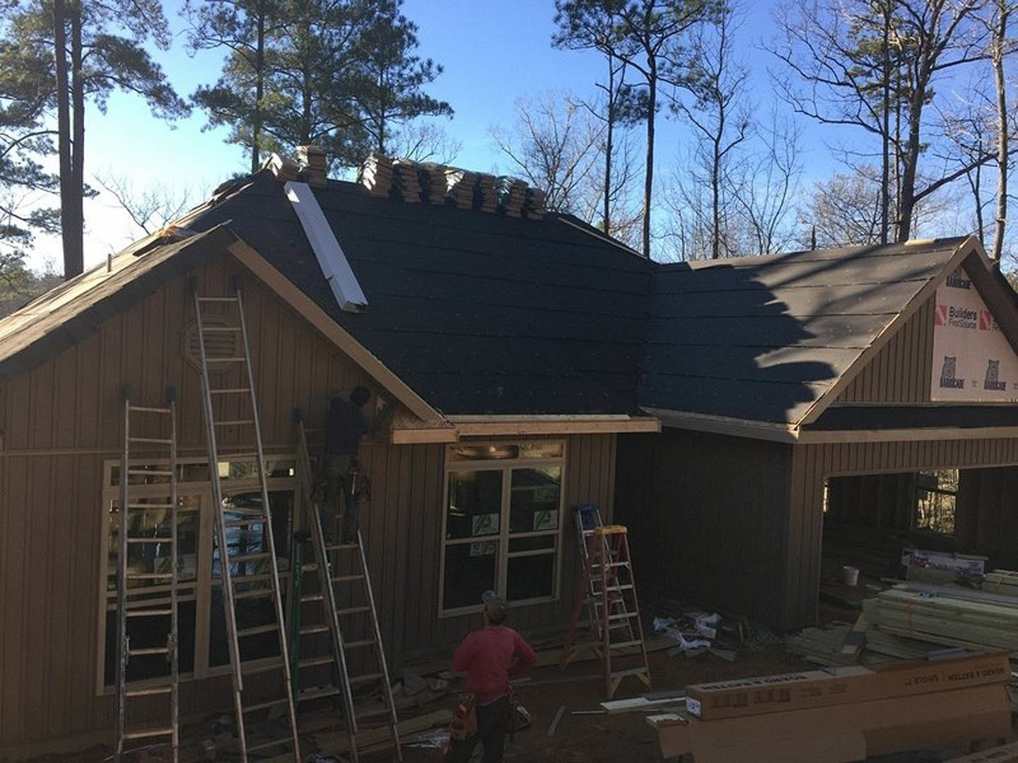 Wood-framed house under construction with multiple ladders, workers in red shirts, exposed siding, and a window displaying a sign; trees and blue sky in background.