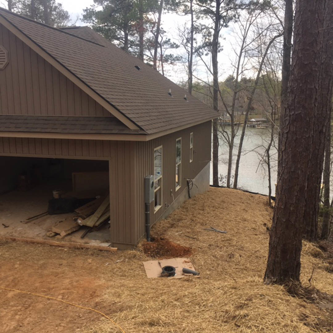 Two-story wood-sided house with attached garage, large windows, and landscaped yard overlooking a lake; mature trees frame the exterior.