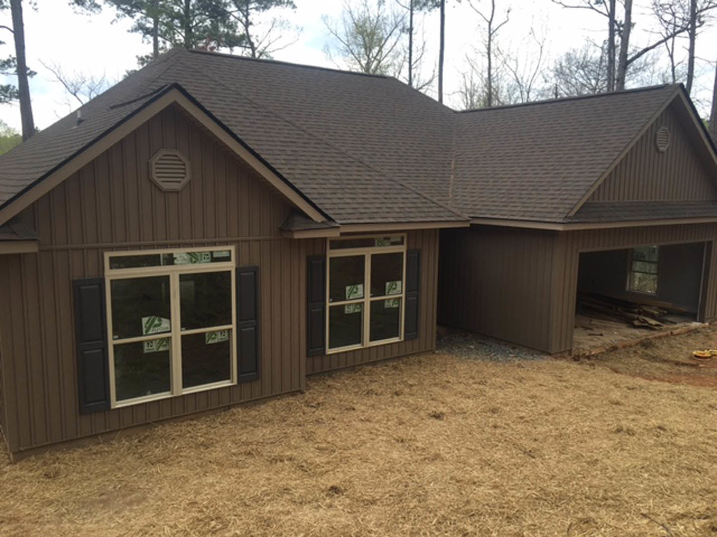 Two-story house with attached garage, brown shingle roof, light siding, front window displaying a sign, wall vent, patchy brown grass in yard, clear sky overhead.