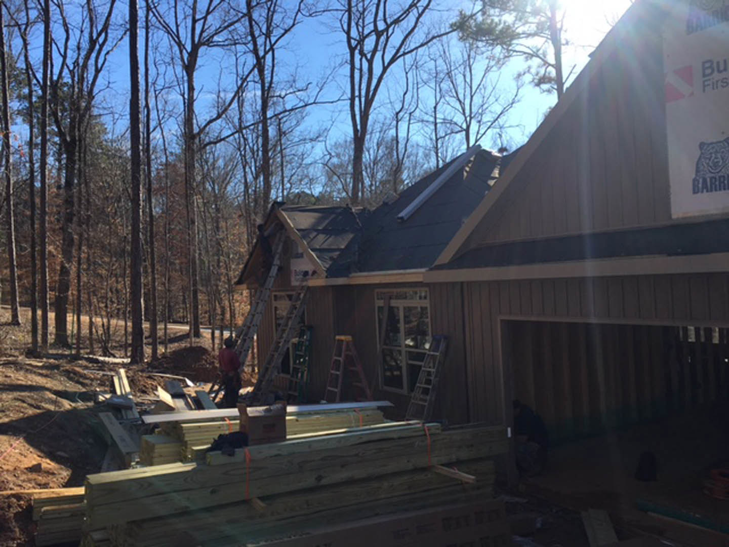 Partially built house framed with exposed lumber, surrounded by leafless trees and winter ground, construction materials stacked nearby