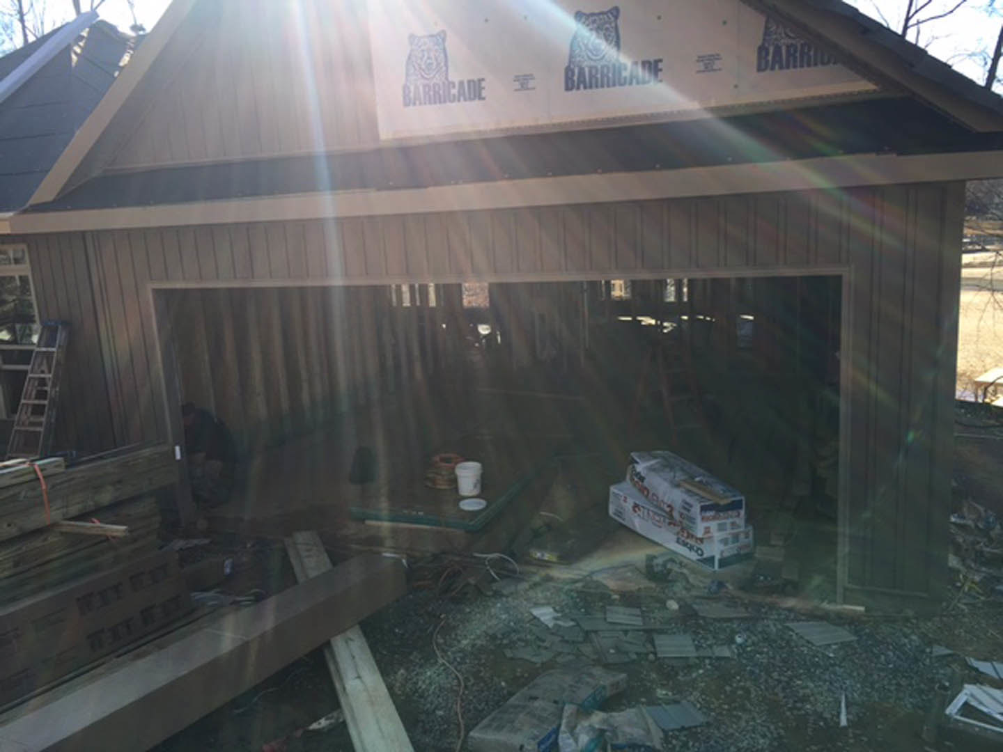 Framed house under construction with sunlight streaming through exposed beams, exterior siding partially installed, ladder leaning against wall, stack of boxes and white sign with