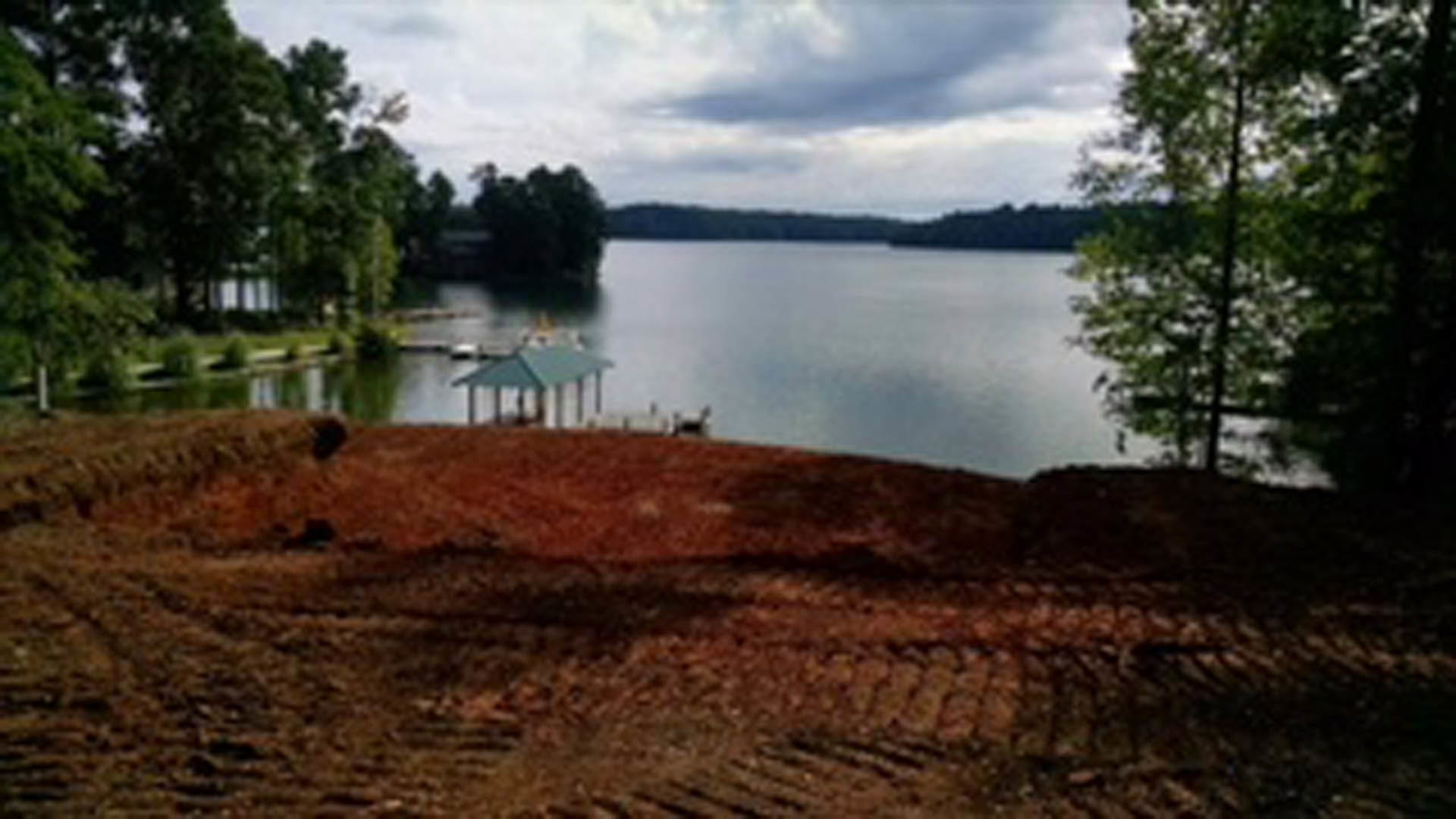 Modern lakeside home with large windows, surrounded by mature trees, overlooking calm water and grassy shoreline under a cloudy sky