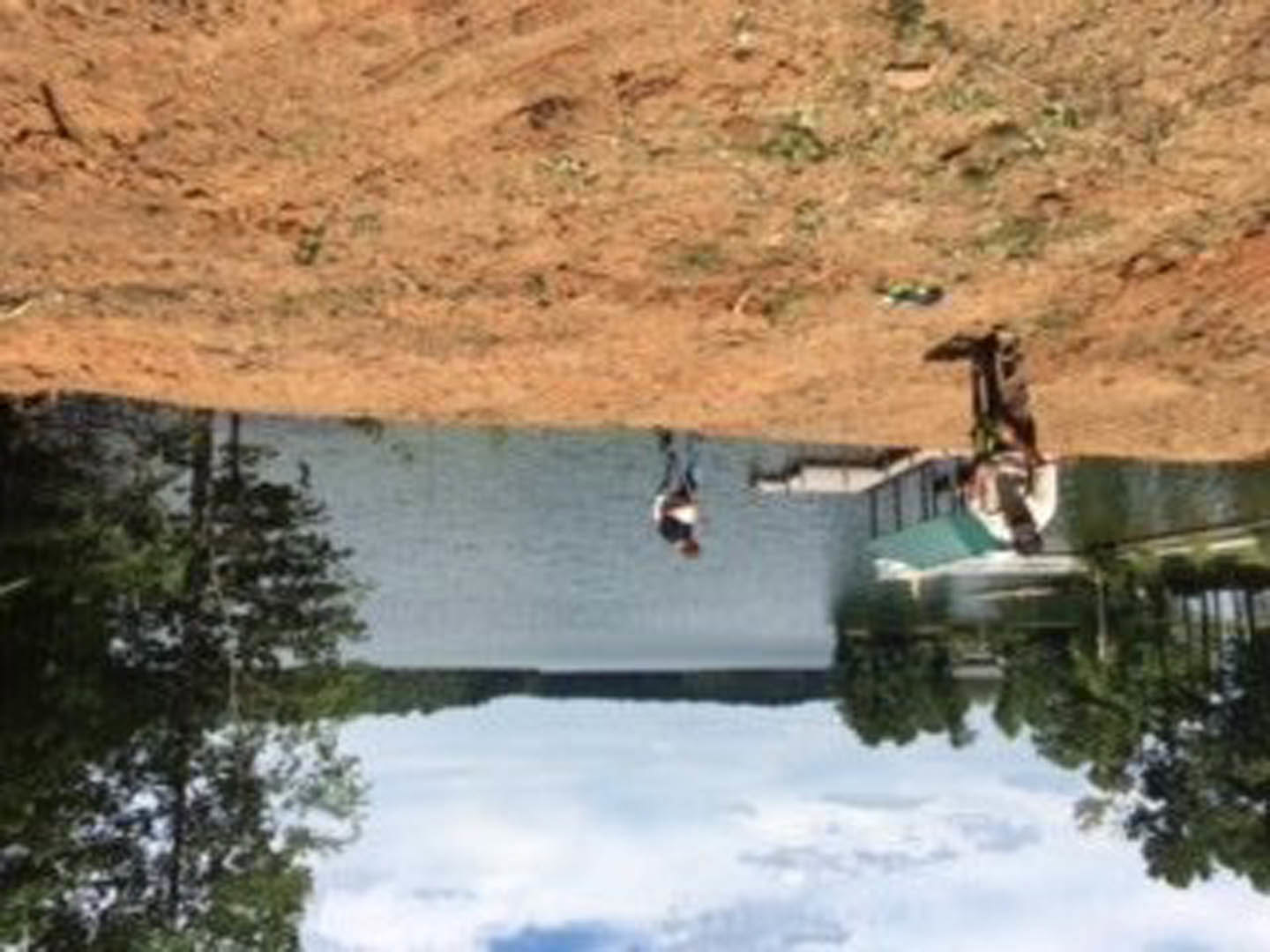 Modern lakeside home with wood siding and large windows overlooking a calm pond, surrounded by trees and mountain landscape, boat docked on water in foreground