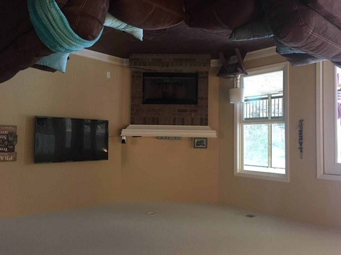 Living room with a stone fireplace, mounted flat-screen television, brown pillow on a gray couch, large window with metal railing, plaster walls, and cabinetry.