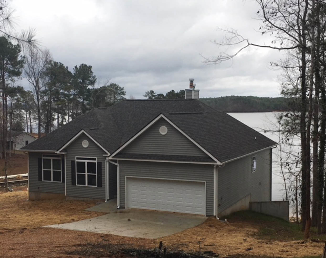 White house with metal roof and attached garage, large windows with white frames, dirt driveway, person standing near lake in background, surrounded by trees under cloudy sky.