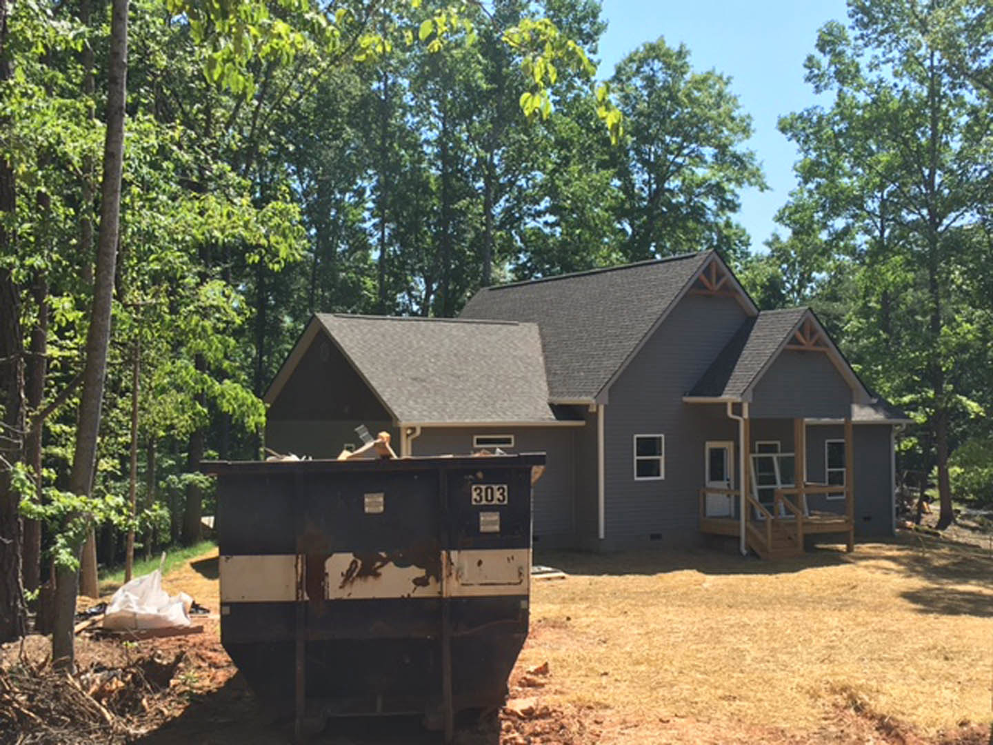 Backyard view of a house with white-framed windows, black and white dumpster, trash can, white plastic bag on dry grass, surrounded by trees and plants