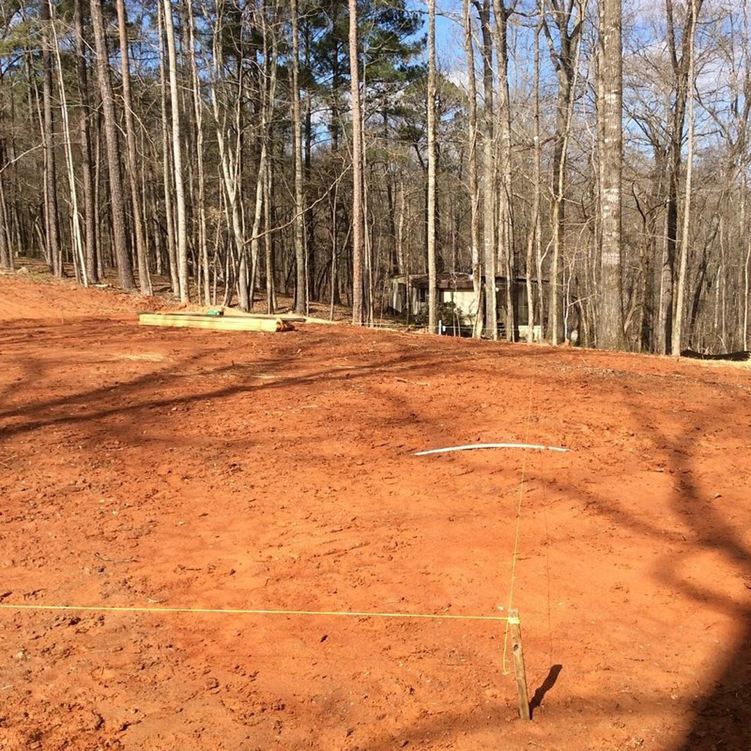 Dirt clearing bordered by tall trees, natural woodland setting, partial shadow cast on soil, custom home partially visible among foliage