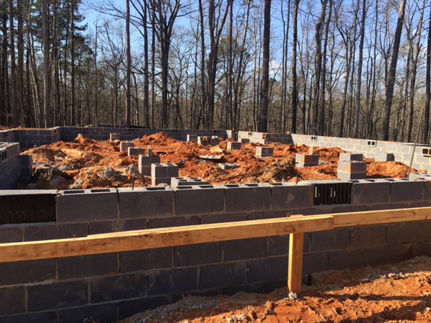 Brick foundation under construction with stacked bricks and exposed dirt, wooden bench beside unfinished brick wall, surrounding trees in background
