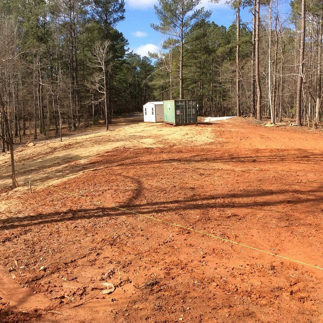 Modern white house with wood siding nestled among tall trees, surrounded by dirt and grass, under a clear sky.