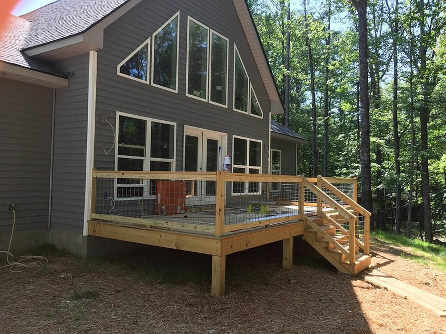 Wooden deck with wire railing and stairs, red door entry, white cord on ground, surrounded by trees and light-colored siding