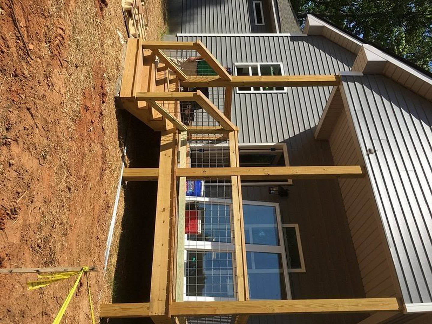 Wooden deck with wire mesh railing, wooden stairs leading to entry, yellow tape wrapped around a beam, black rectangular object near the porch, window framed by wooden beams