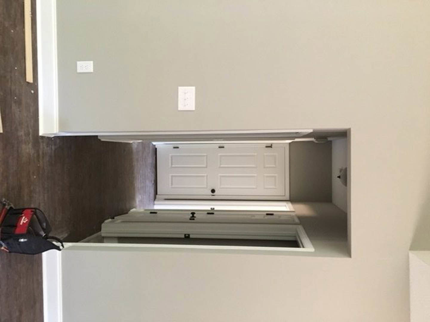 White hallway with paneled door featuring black knobs, light walls, and visible kitchen cabinetry and countertop in the background; person holding a black and red bag near a white