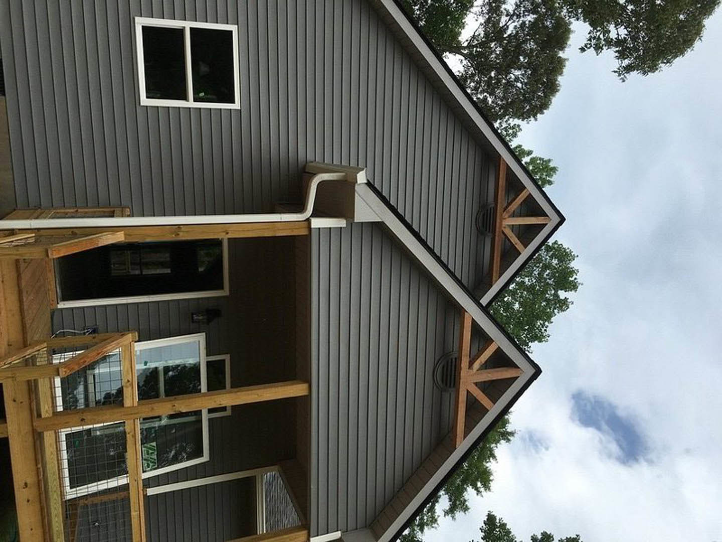 Two-story house with light wooden siding, white-framed windows, covered front porch, gray shingle roof, leafy tree in foreground, and cat enclosure attached to side