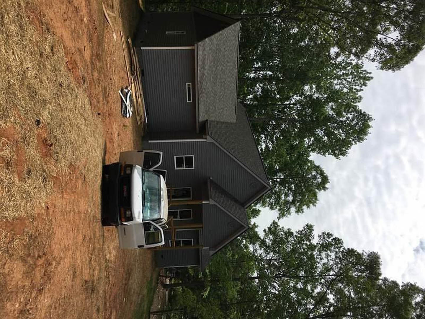 Two-story custom home with light siding, white-framed windows, and a white van parked on the gravel driveway; mature leafy tree in front yard, partly cloudy sky overhead.
