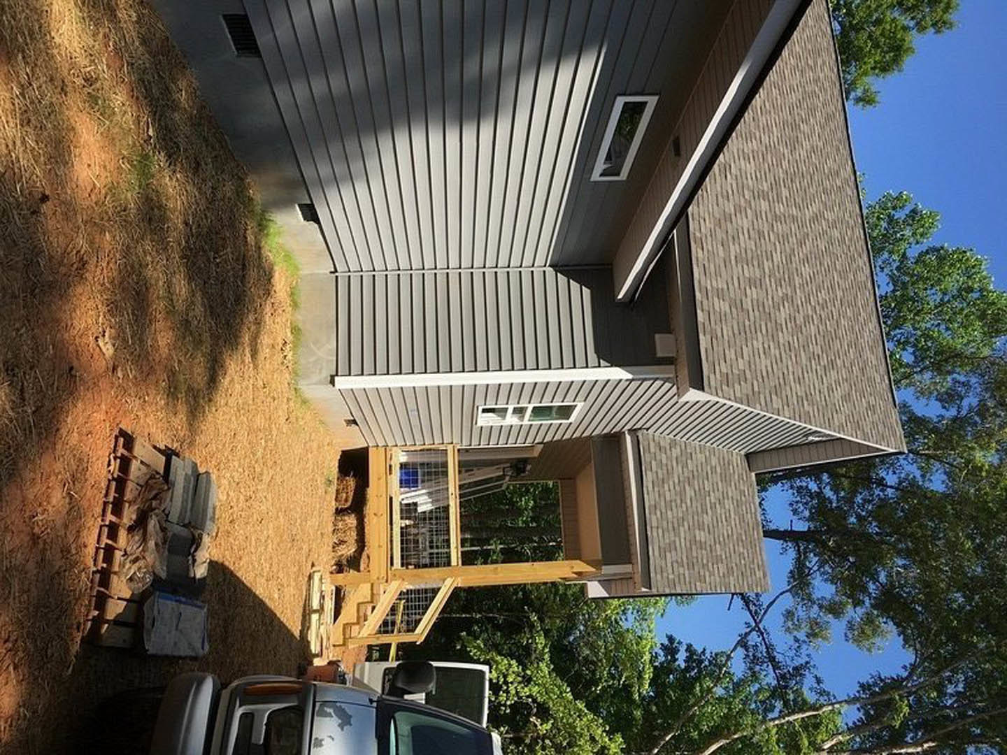 Two-story house with white siding, fenced yard, parked pickup truck in driveway, large windows, wooden pallet on ground, mature trees, blue sky