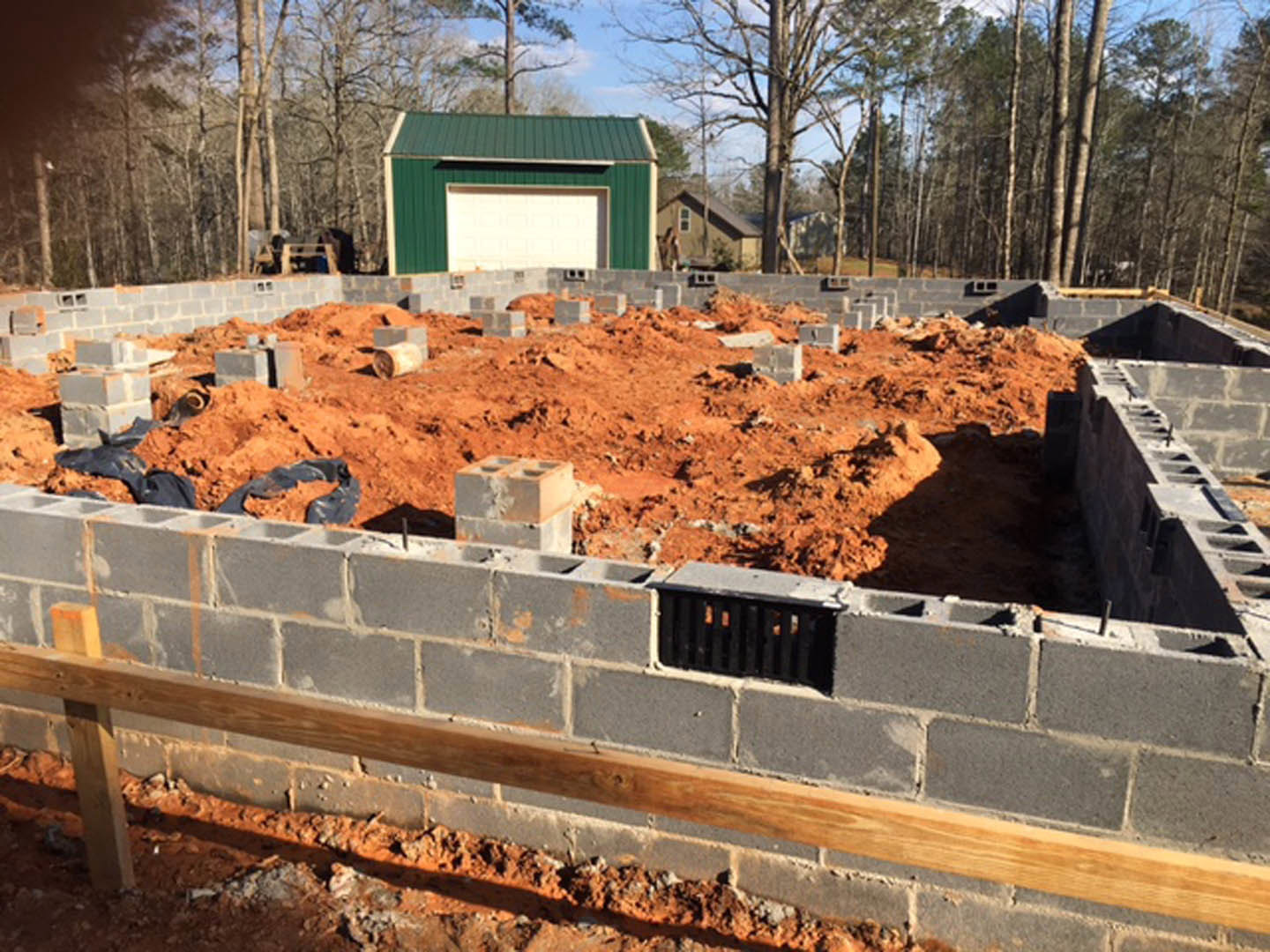 Concrete foundation with exposed brick wall, metal grate, green garage with white door, pile of bricks and dirt, trees and sky in background