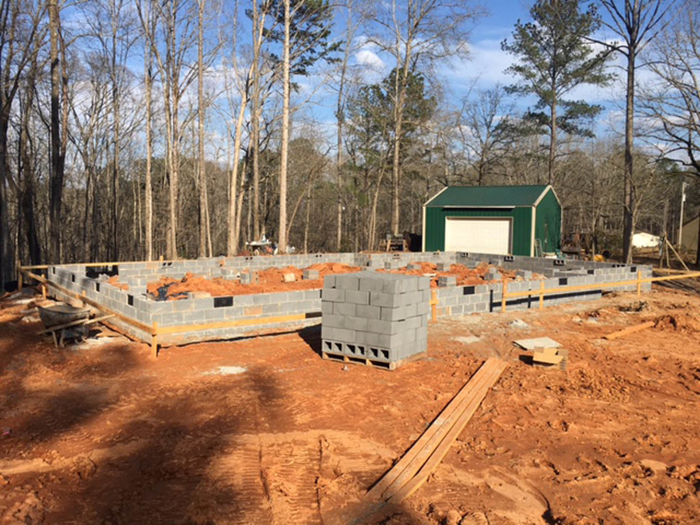 Green garage with white roof beside partially built brick walls, stacks of grey bricks and wooden planks on soil, surrounded by trees under a clear sky
