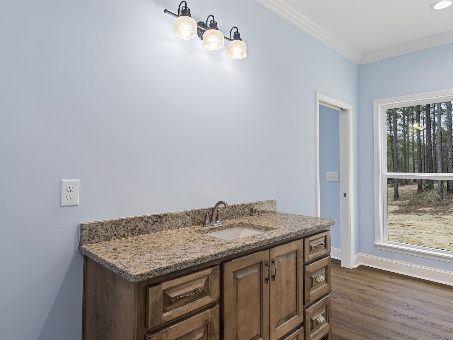 Bathroom with marble countertop, undermount sink, white cabinetry, three-light fixture above mirror, white electrical outlet, window overlooking trees, light-colored tile walls