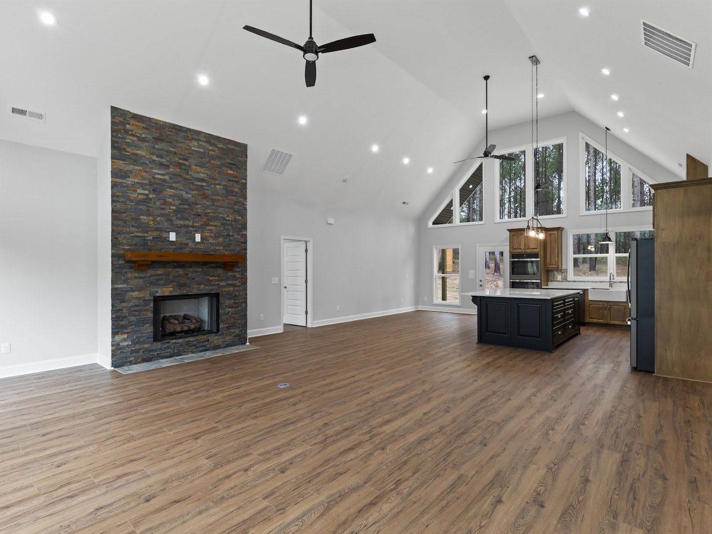 Spacious living area with hardwood floors, stone fireplace featuring a wood mantel and basket of logs, black kitchen island with white countertops, ceiling fan with light fixture