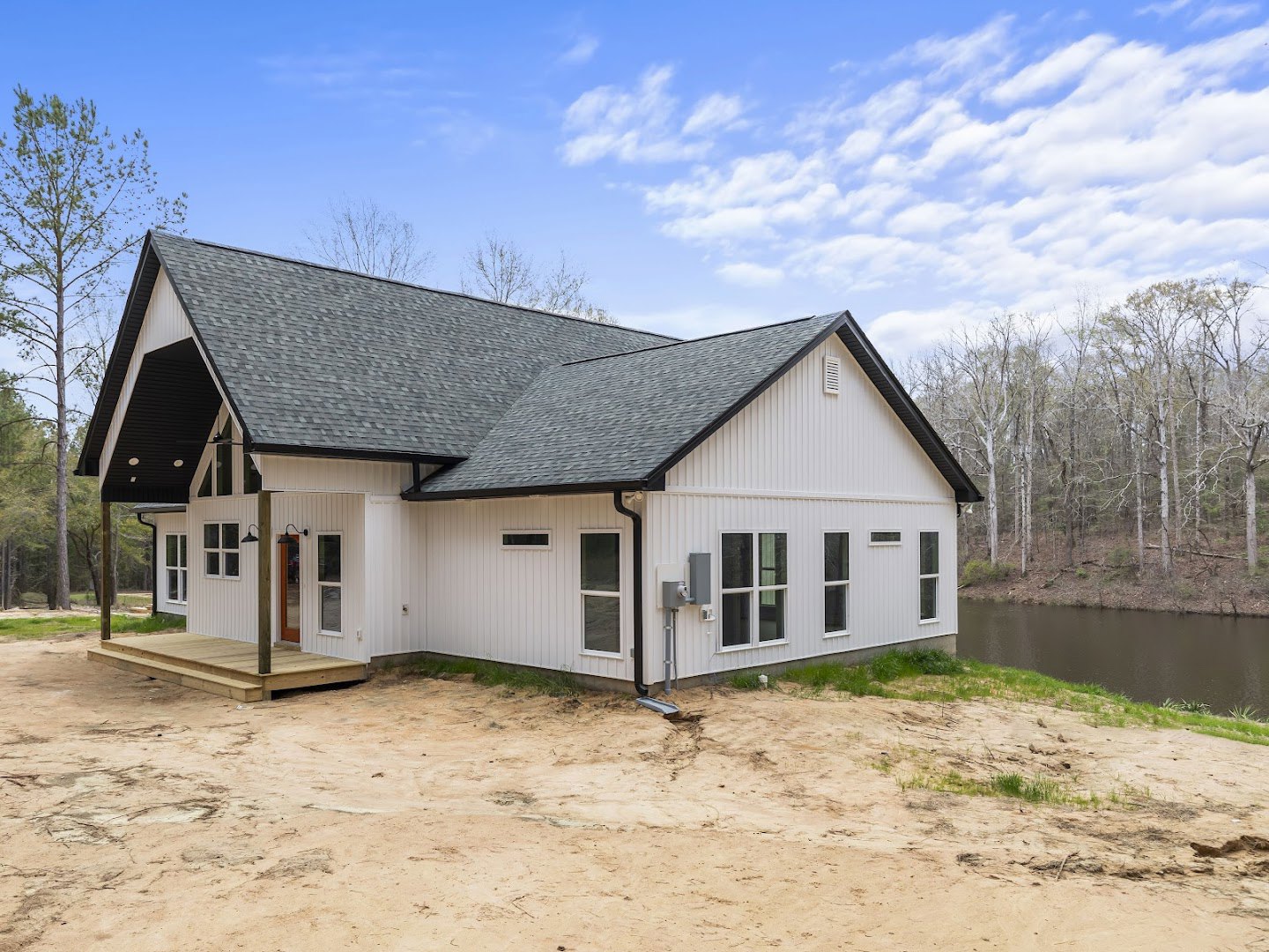 White cottage-style house with wood front door, white-framed windows, and drainpipe set in bare dirt; leafless tree stands nearby, lake and Robert Frost Farm visible in the